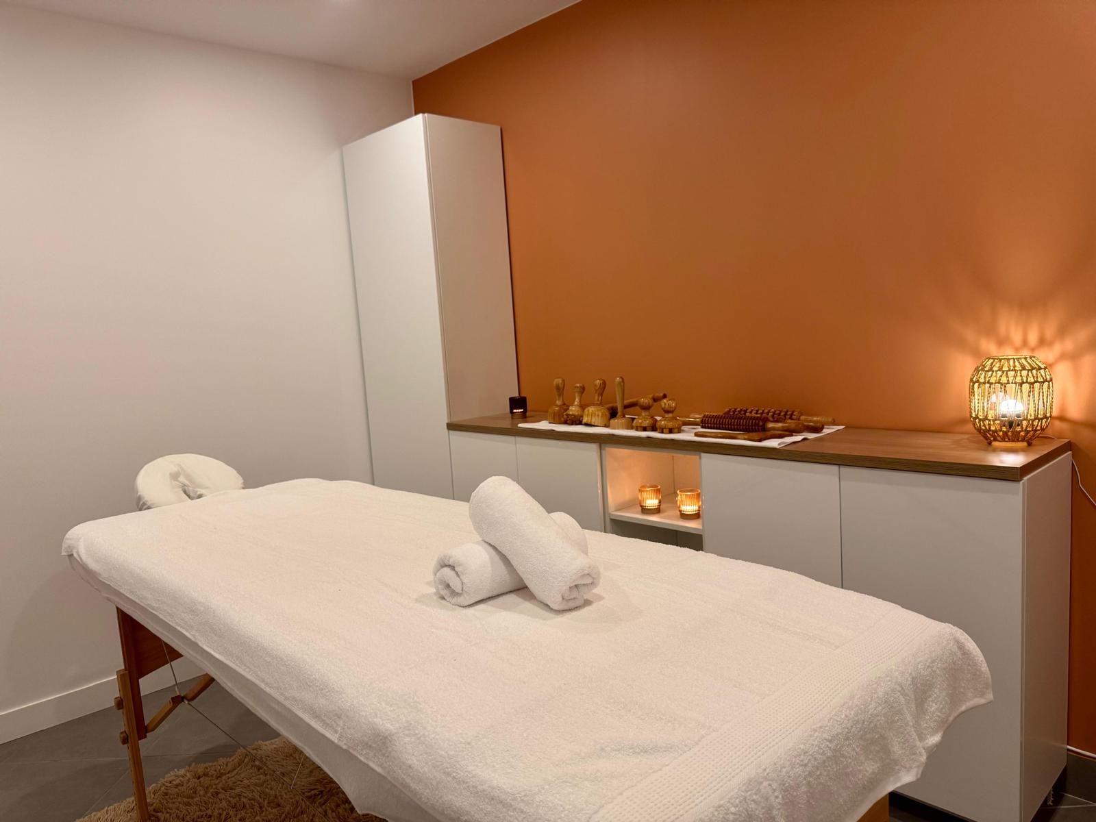 Cabine de soin : table de massage, serviettes roulées, outils de madérothérapie en bois et mur orange.