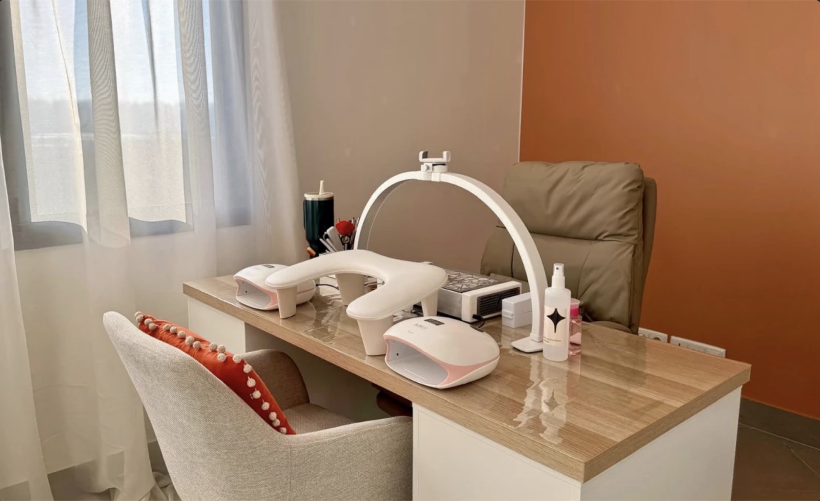 Poste de manucure près de la fenêtre : table en bois clair, lampe en arc, repose-mains et lampes de séchage, fauteuil beige devant un mur orange.